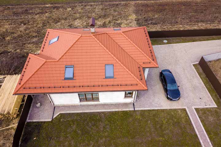 Aerial top view of house metal shingle roof with attic windows and black car on paved yard.